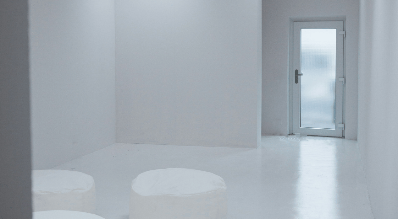 A white, glass door in a completely bare, white clinic looking room. There are two stools in the middle of the room.