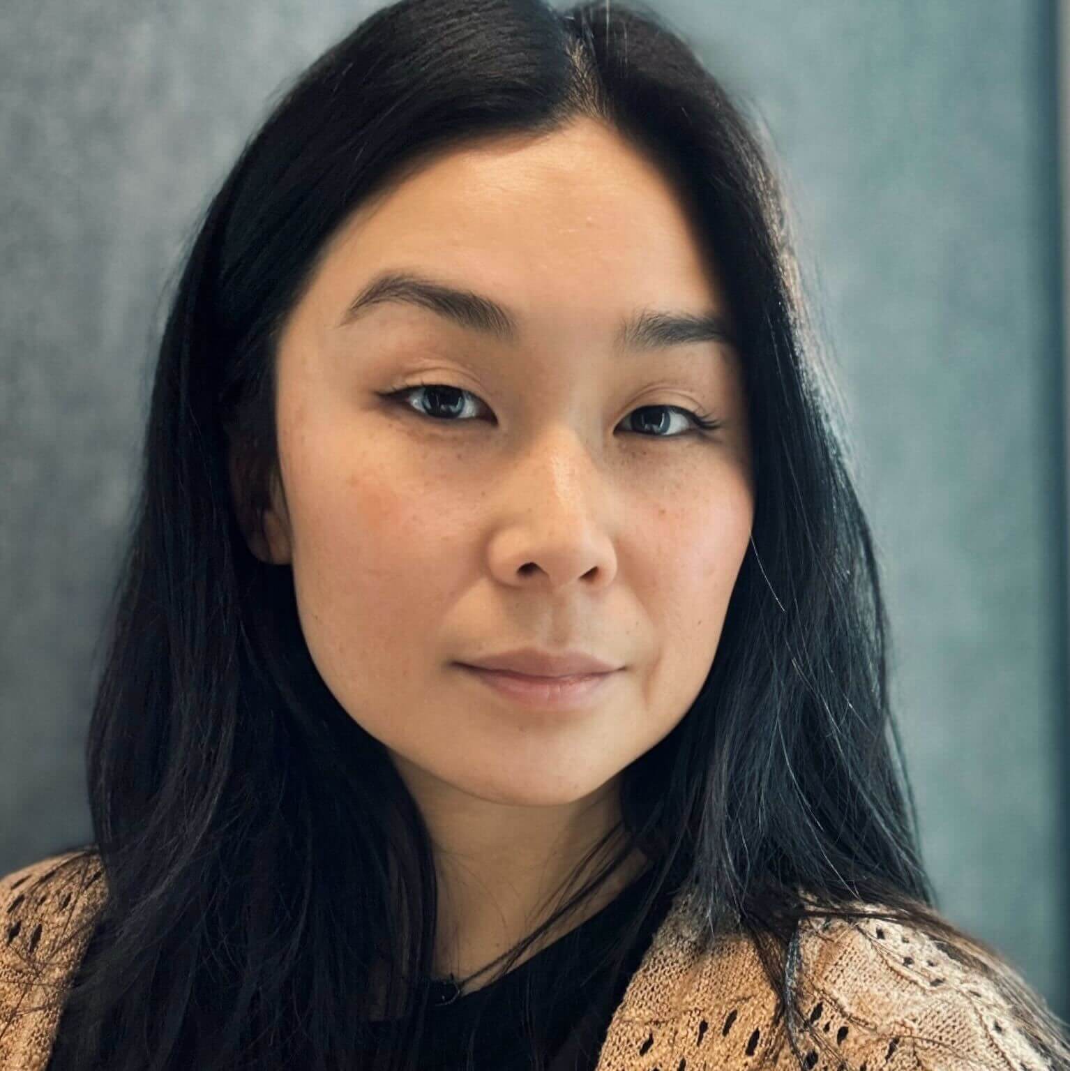 A woman with long, dark hair is wearing a black top and a light sweater, set against a blurred background.