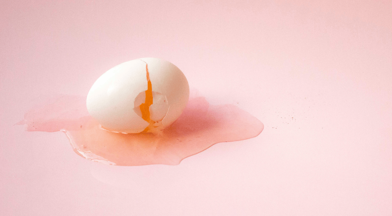 A cracked white egg on a pale pink background with the yolk spilled around it.