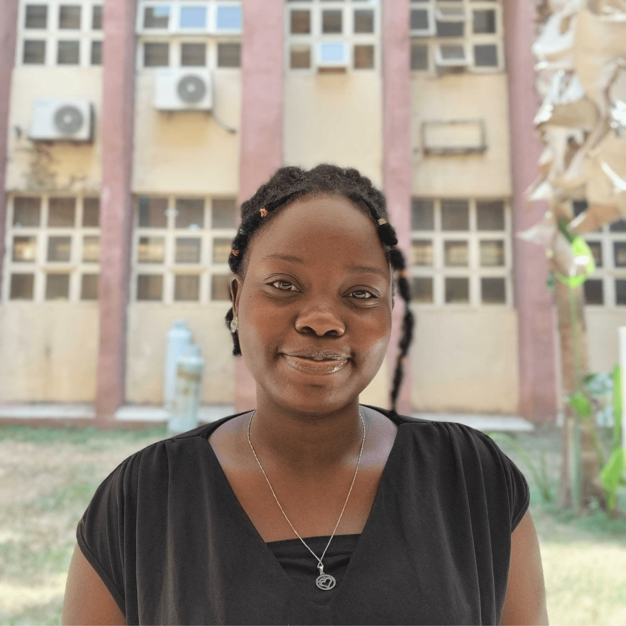 A woman wearing a black top stands outdoors, with a building featuring large windows and air conditioning units in the background.