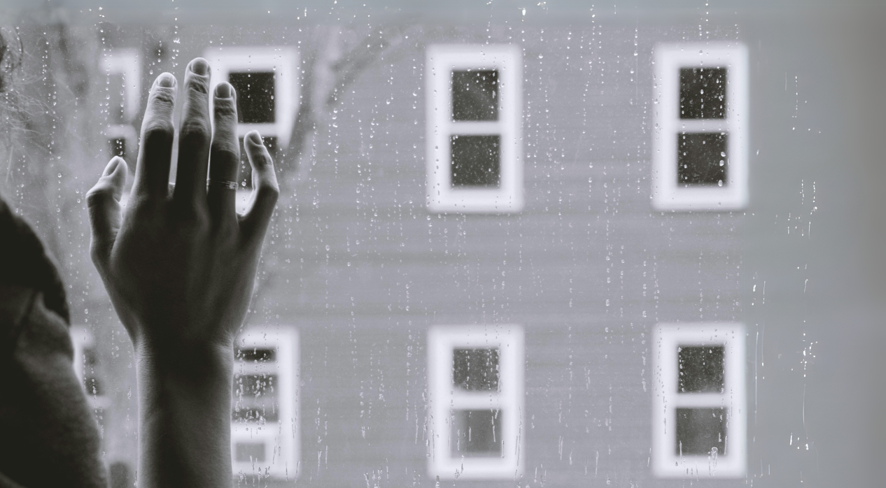 Hand with a ring against a rain-speckled window with a building in the background.