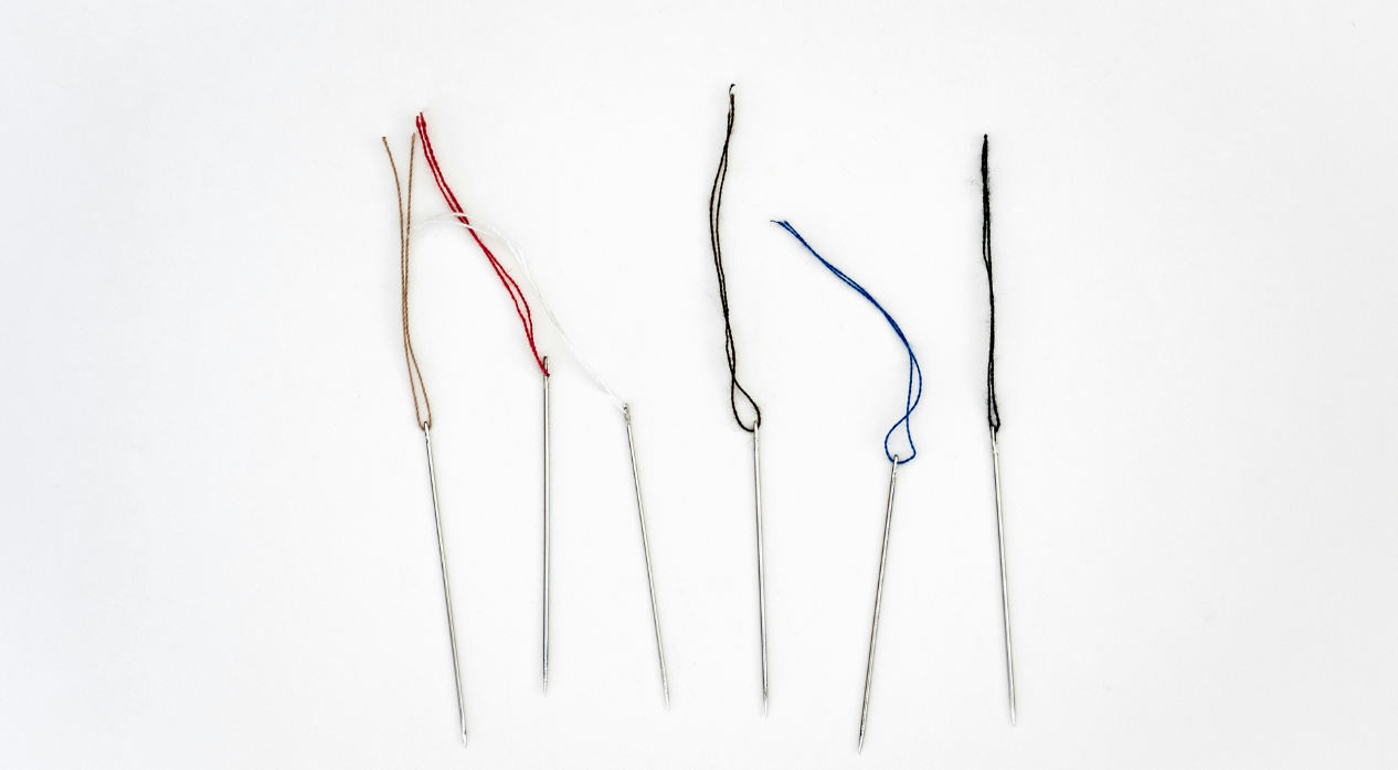 Five needles threaded with tan, red, white, blue, and black threads on a white background.