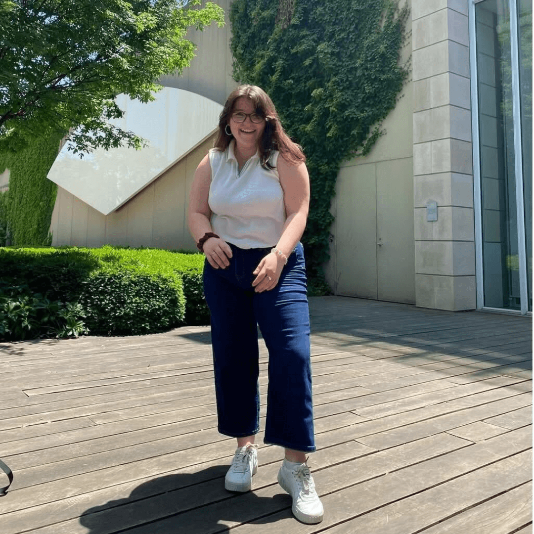 A woman stands on a wooden deck surrounded by green foliage, wearing a white top and navy pants, under a clear blue sky.