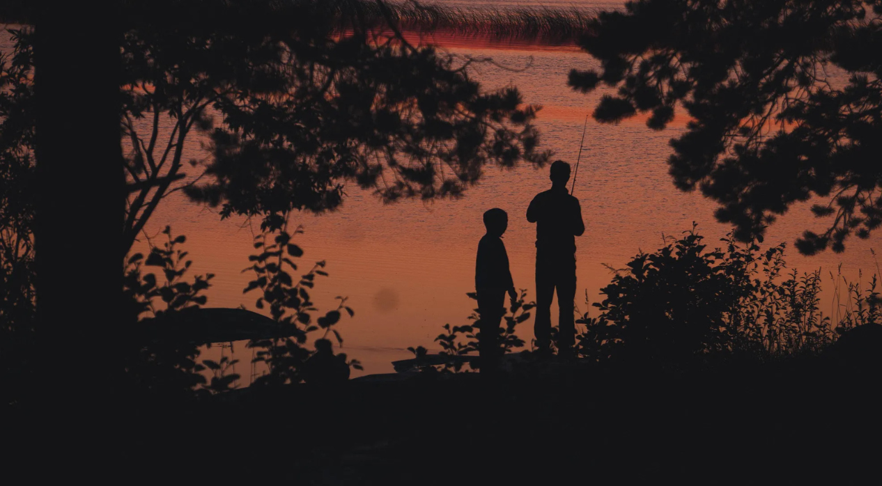 Silhouettes of two people fishing by a lake at sunset, surrounded by trees.