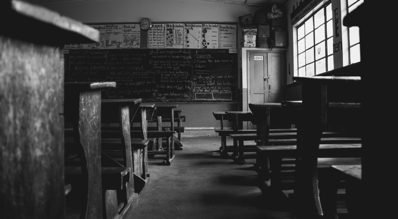 Black and white photo of an empty classroom with wooden desks facing a chalkboard full of notes.