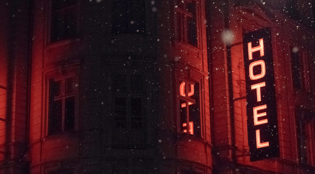 A building exterior at night with glowing red neon "HOTEL" sign and falling snow.