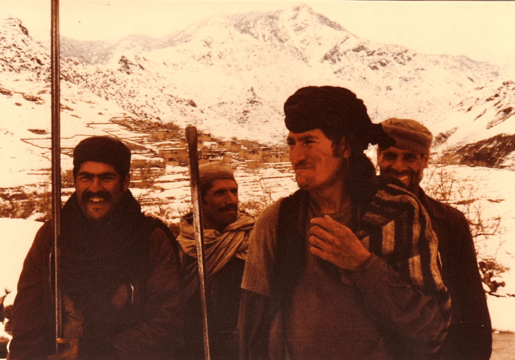 Four men in winter clothing in a snowy mountain landscape with a village in the background.