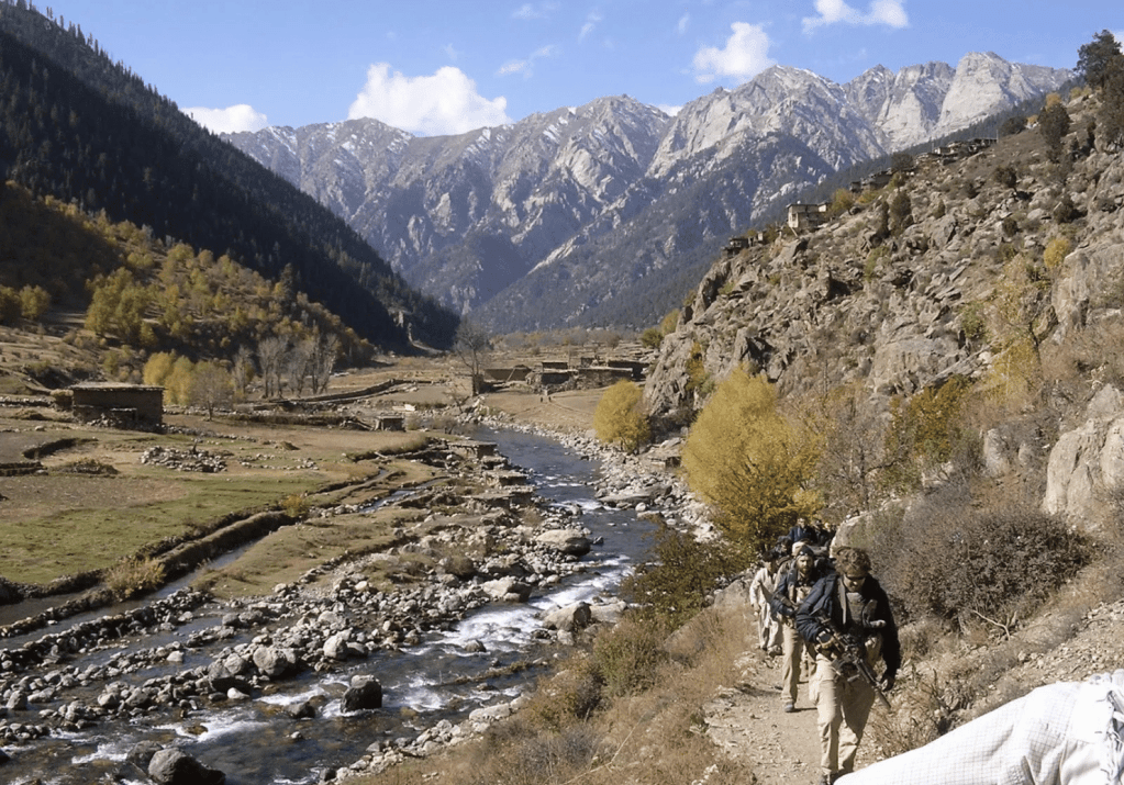 A group of people walking along a trail in a mountainous valley with a river, trees, and snow-capped peaks.