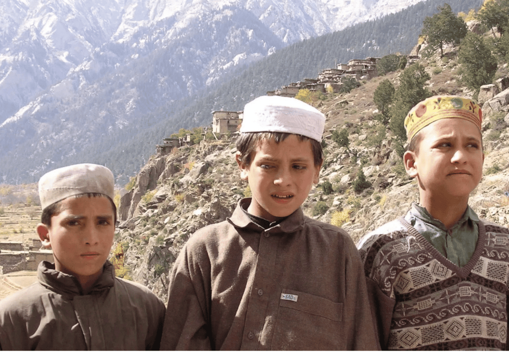 Three boys wearing caps stand in front of a mountainous landscape.
