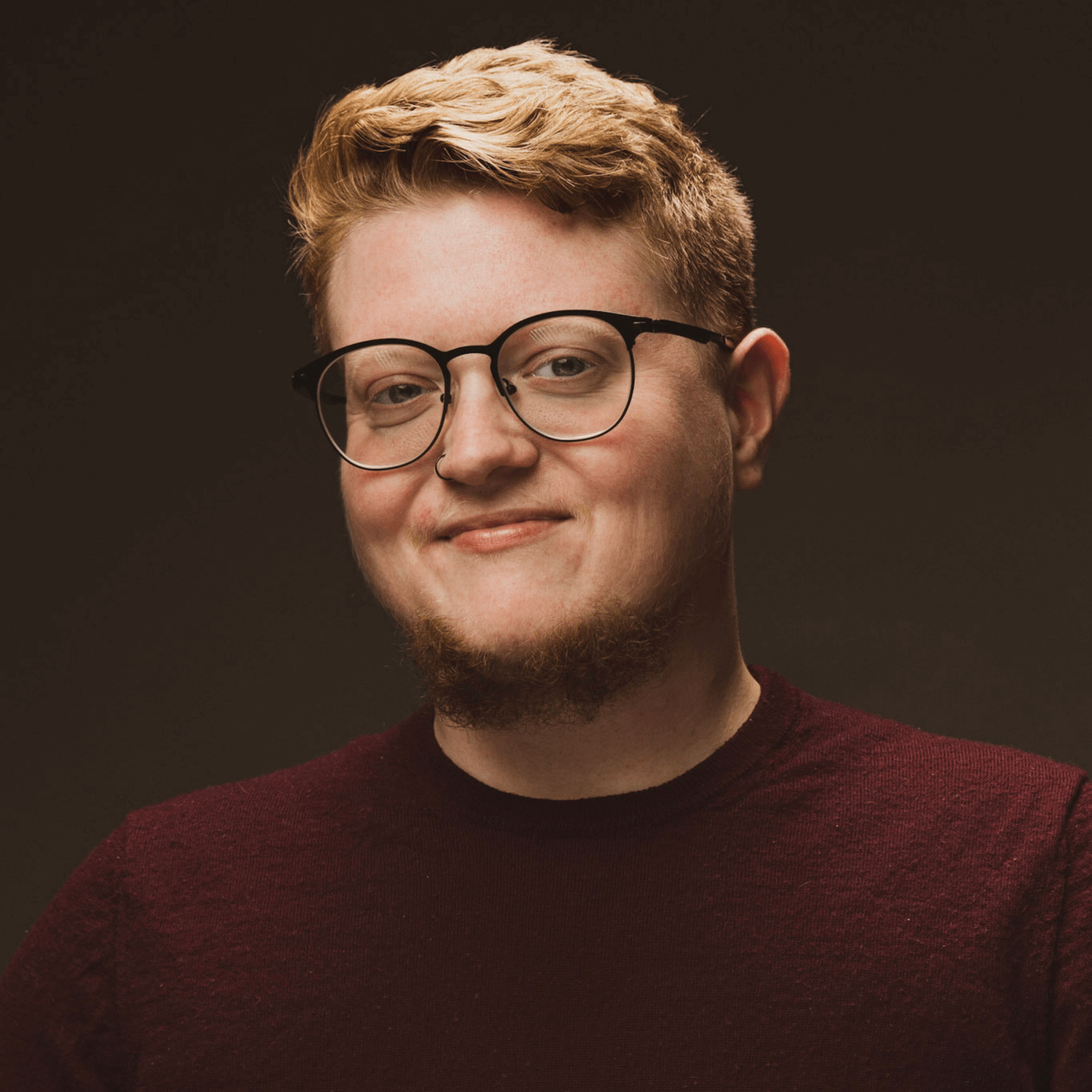 A man with glasses and light blonde hair wears a dark maroon sweater, set against a dark background.