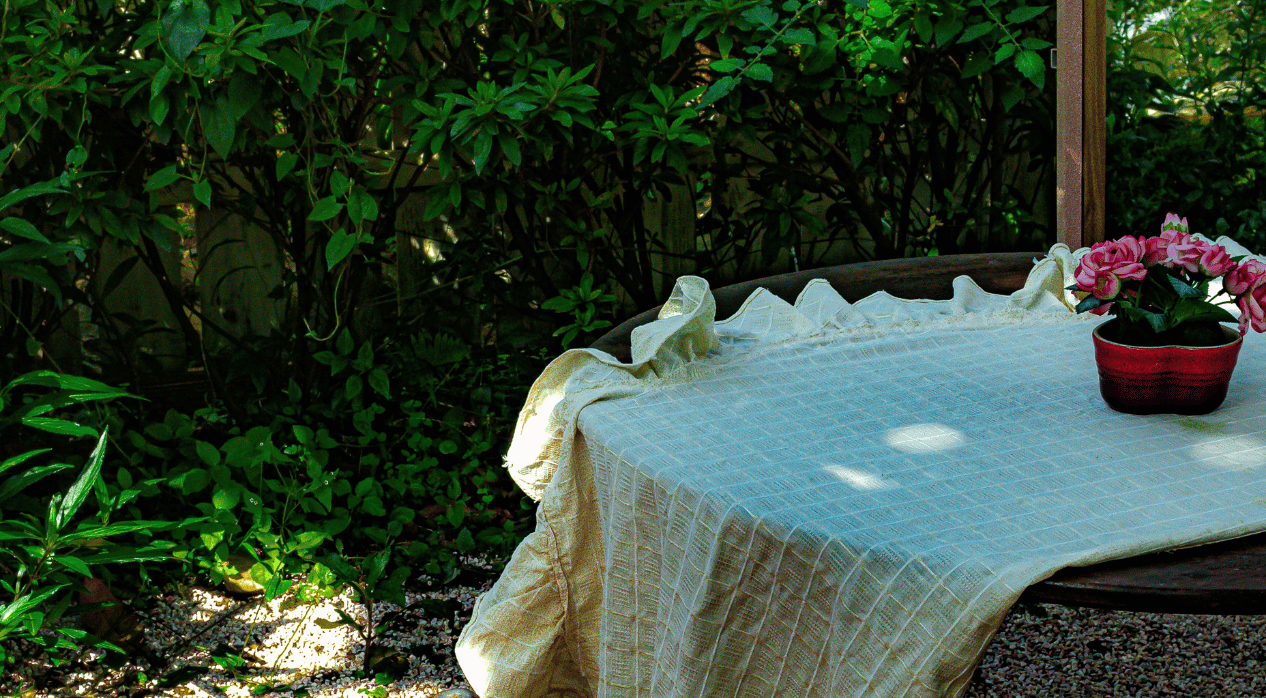 A wooden table outdoors is covered with a cream-colored tablecloth and holds a red pot with pink flowers, surrounded by green bushes and plants.